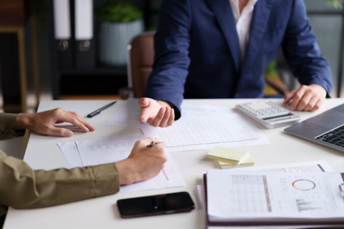 Two individuals engaged in a discussion at a desk, with a focus on their hands and a document. A laptop displaying charts and graphs is visible in the background